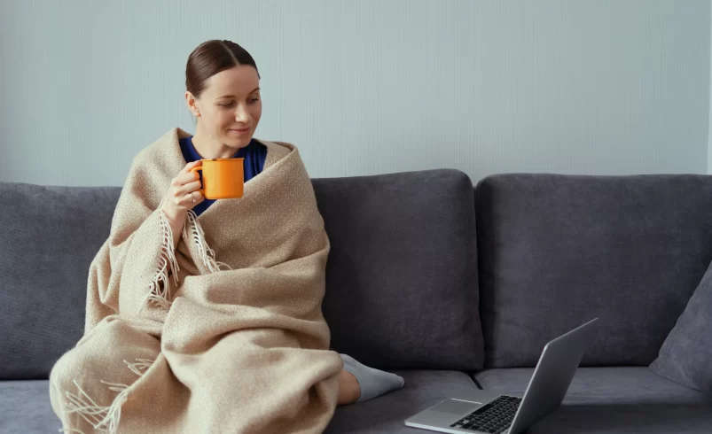 Žena sedí uvelebená na gauči s elektrickou vyhřívanou dekou